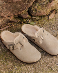 Women's Birkenstock Boston Shearling Clogs in Taupe Suede with natural cork sole and plush lining, styled on rugged stone surface