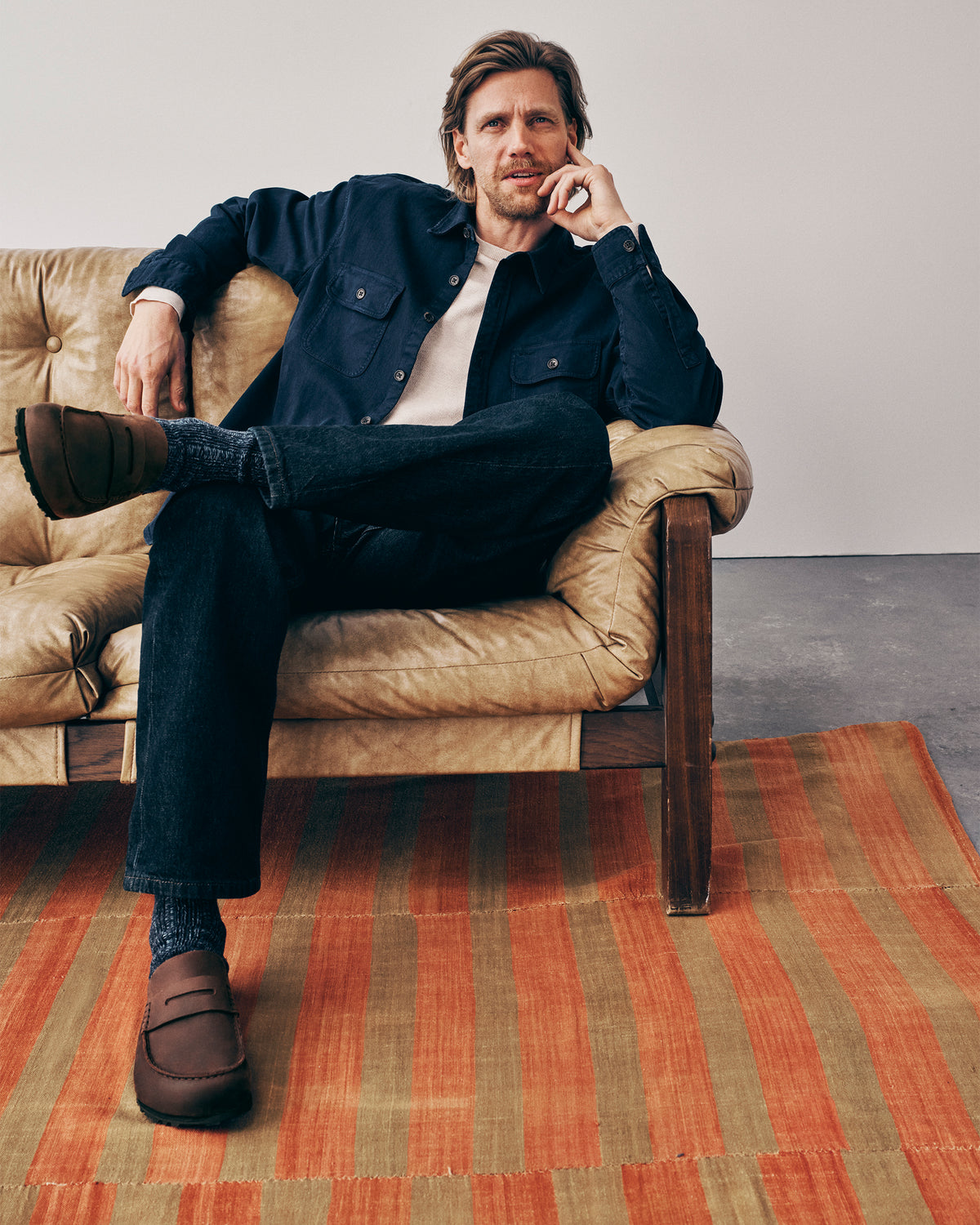 Man sitting on a brown leather chair wearing a navy jacket, dark jeans, and Birkenstock Naples Wrapped Clogs in Habana Oiled Leather.