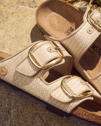Beige sandals with gold buckles on a textured surface. Birkenstock Arizona Big Buckle Sandal in Raffia Naturale Textile