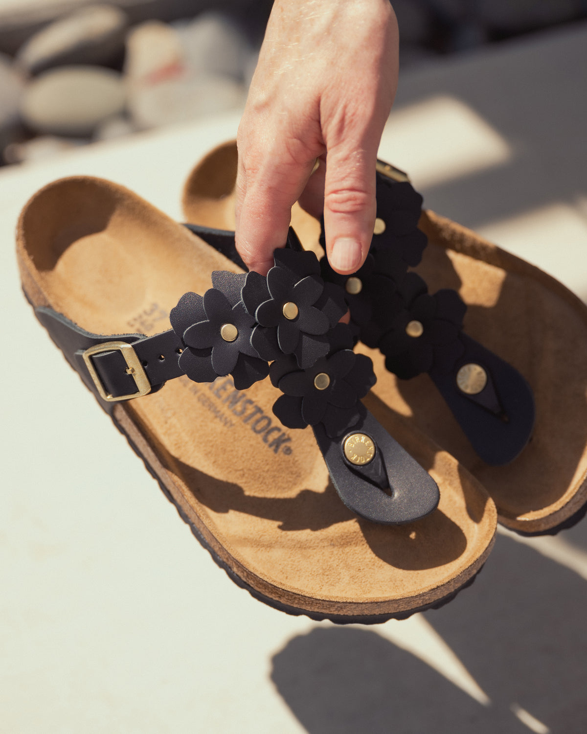 Person holding a Birkenstock Gizeh Flowers Sandal in Black Leather with black floral applique straps on a blurred background