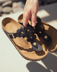 Person holding a Birkenstock Gizeh Flowers Sandal in Black Leather with black floral applique straps on a blurred background