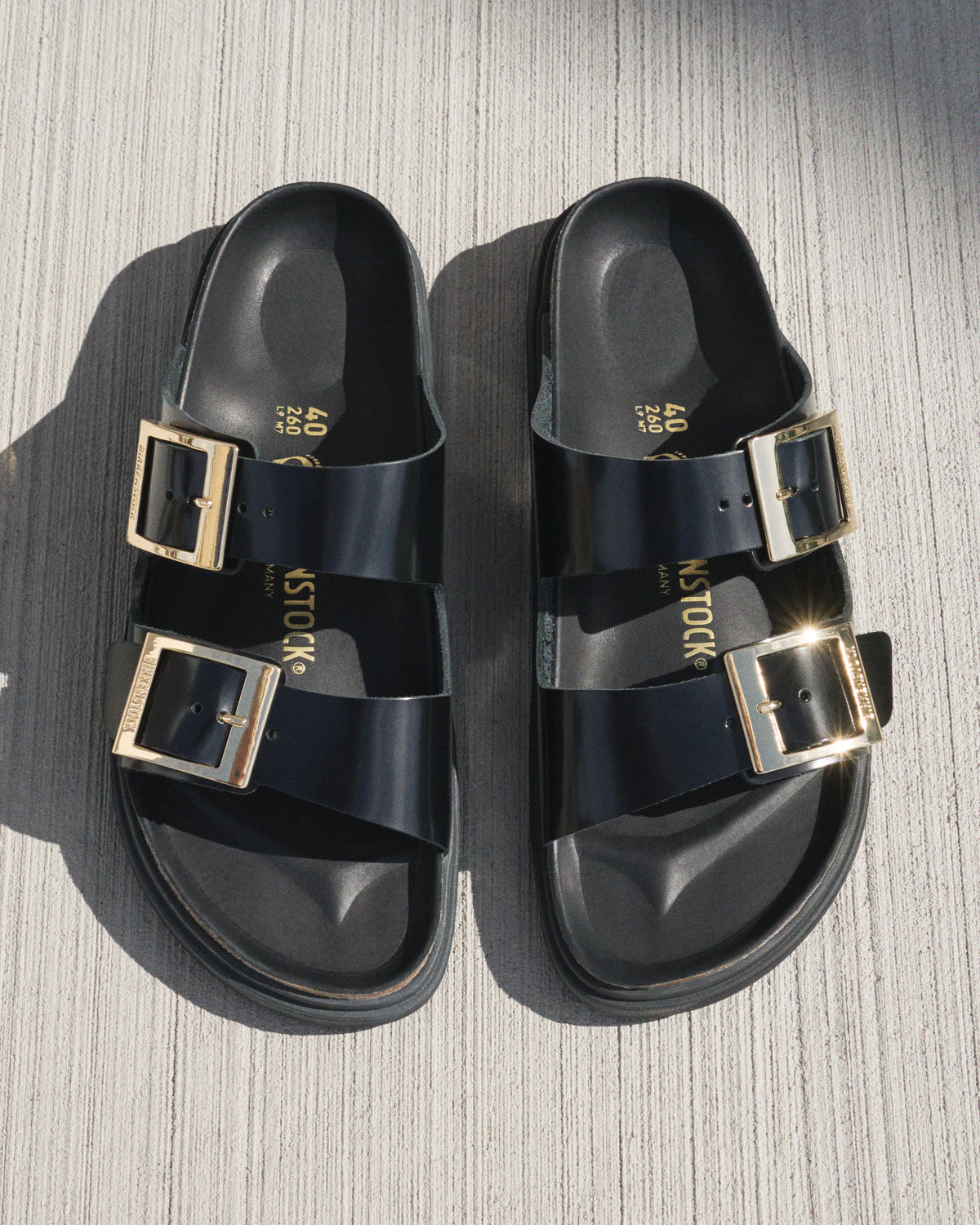 Black Birk sandals with gold droplet buckles on a textured surface