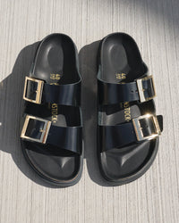 Black Birk sandals with gold droplet buckles on a textured surface