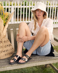 Woman sitting on a wooden bench wearing a white shirt, denim shorts, and black patent leather two-strap Birkenstock sandals with a straw bag next to her.