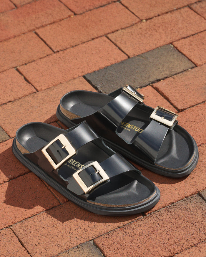 Luxurious black high shine Birkenstock Arizona Droplet Buckle sandals with gold buckles on a brick surface