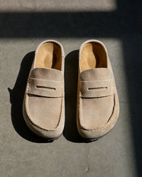 A pair of beige suede Birkenstock Naples Wrapped Clog in Taupe Suede, displayed on a dark gray concrete surface under direct sunlight with sharp shadows.