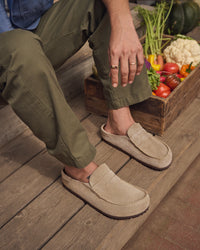 Person wearing Birkenstock Naples Wrapped Clogs in Taupe Suede on a wooden floor with a box of vegetables in the background