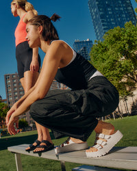Two women in athletic wear sitting on a bench with cityscape in the background. Woman in front wearing the Birkenstock EVA Florida Flex Platform Sandals in Eggshell and woman in back wearing the Birkenstock EVA Arizona sandals in Black