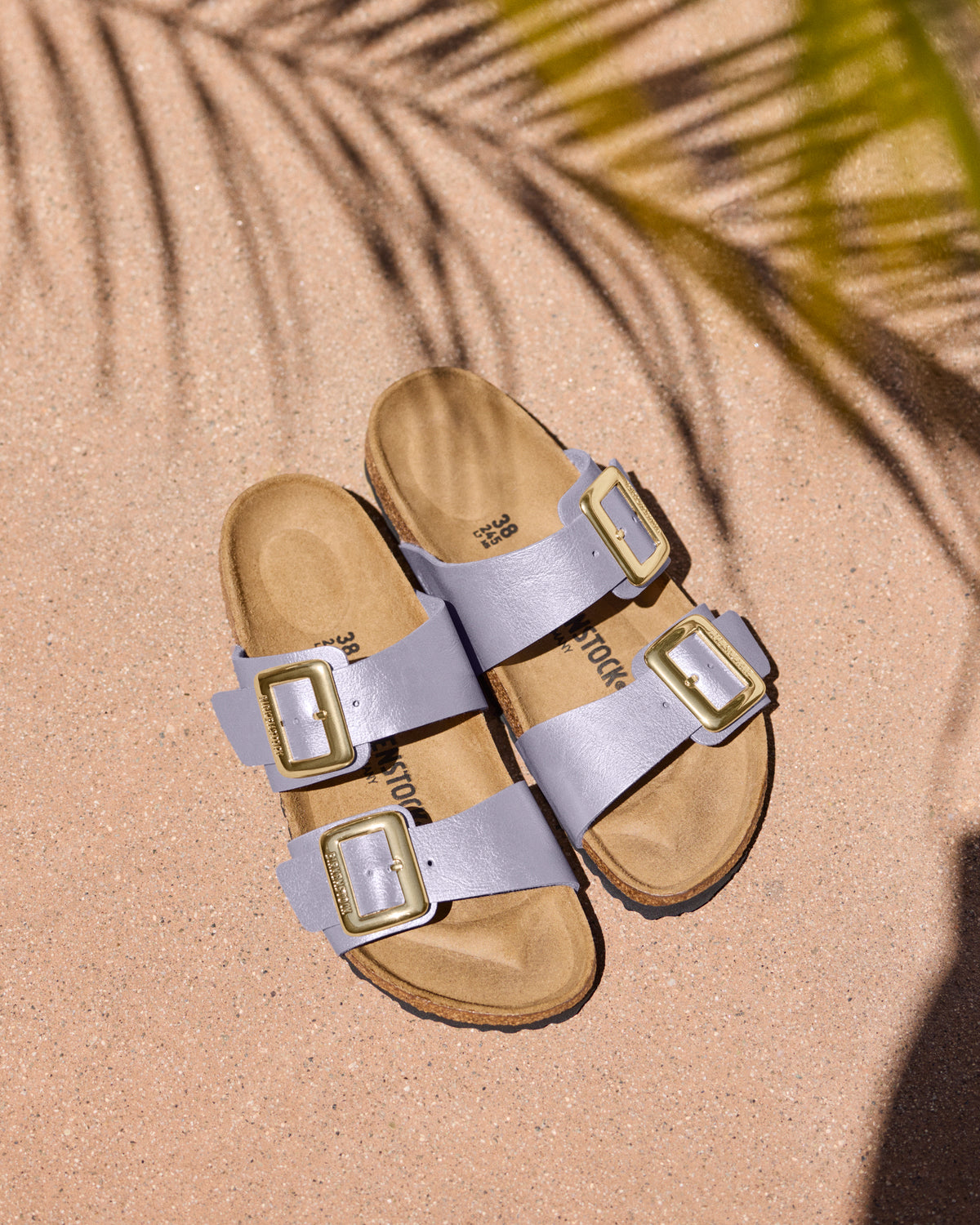 Pair of Birkenstock Sydney Luxe Buckle Sandals in Graceful Orchid Purple with tan soles on a gritty background