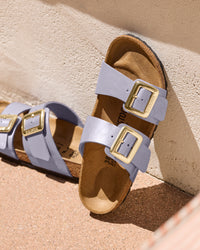 Pair of Birkenstock Sydney Luxe Buckle Sandals in Graceful Orchid Purple with gold buckles on a concrete surface