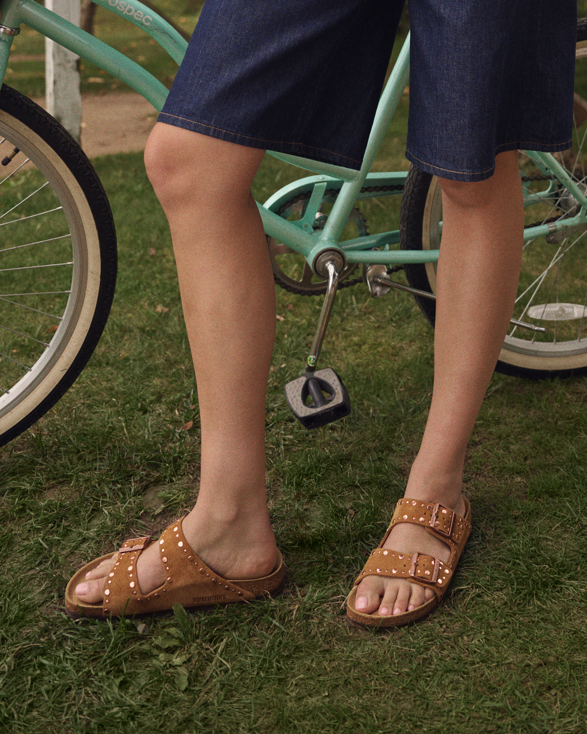 Person wearing Mink brown Birkenstock Arizona double-strap sandals with studs standing on grass next to a teal bicycle.