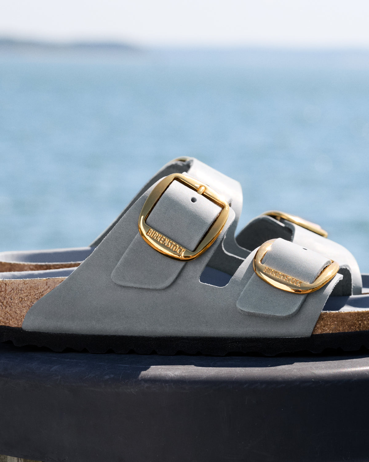 Birkenstock Arizona Big Buckle sandals in blue leather called "Basalt" with two straps and two gold buckles with "BIRKENSTOCK" on them, pictured on a dark surface with a blurred ocean background