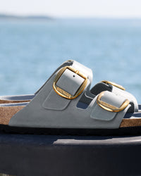 Birkenstock Arizona Big Buckle sandals in blue leather called "Basalt" with two straps and two gold buckles with "BIRKENSTOCK" on them, pictured on a dark surface with a blurred ocean background