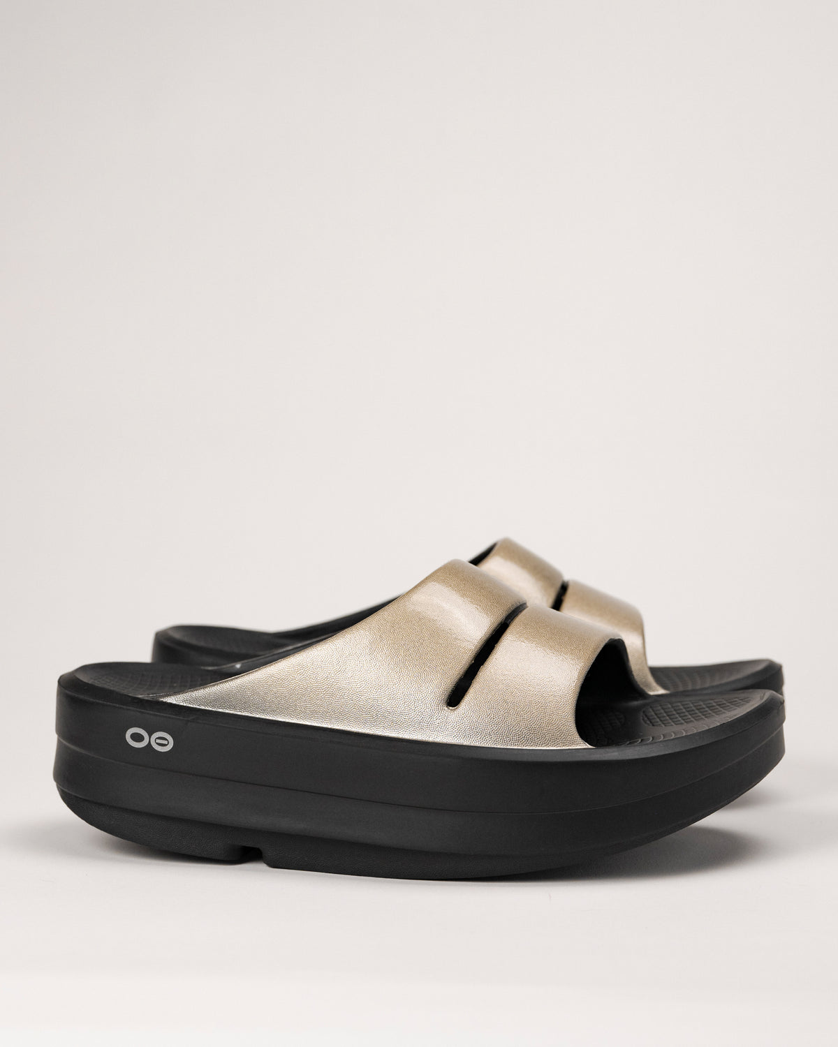 Side view of OOFOS OOmega OOahh Luxe Slide in Latte with golden shimmer upper and black platform sole