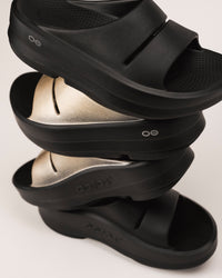 Stack of black and Latte golden OOFOS platform sandals on a light background