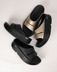 Black and gold OOFOS platform recovery slide sandals on a light background