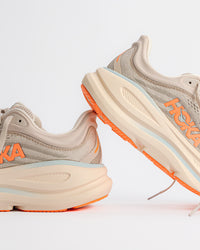 A pair of Women’s HOKA Bondi 9 – Max-Cushioned Running Shoe in River Rock Grey / Mineral Ore - 1162012-RRYM in light beige and gray with bright orange accents and a thick cream-colored sole, shown against a clean white background.