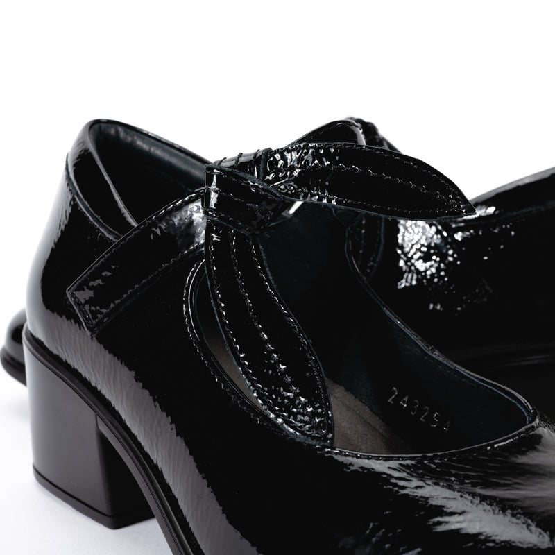 Close-up of the Nobility Mary Jane shoe in glossy black patent leather, highlighting the removable bow on it's hook and loop strap.