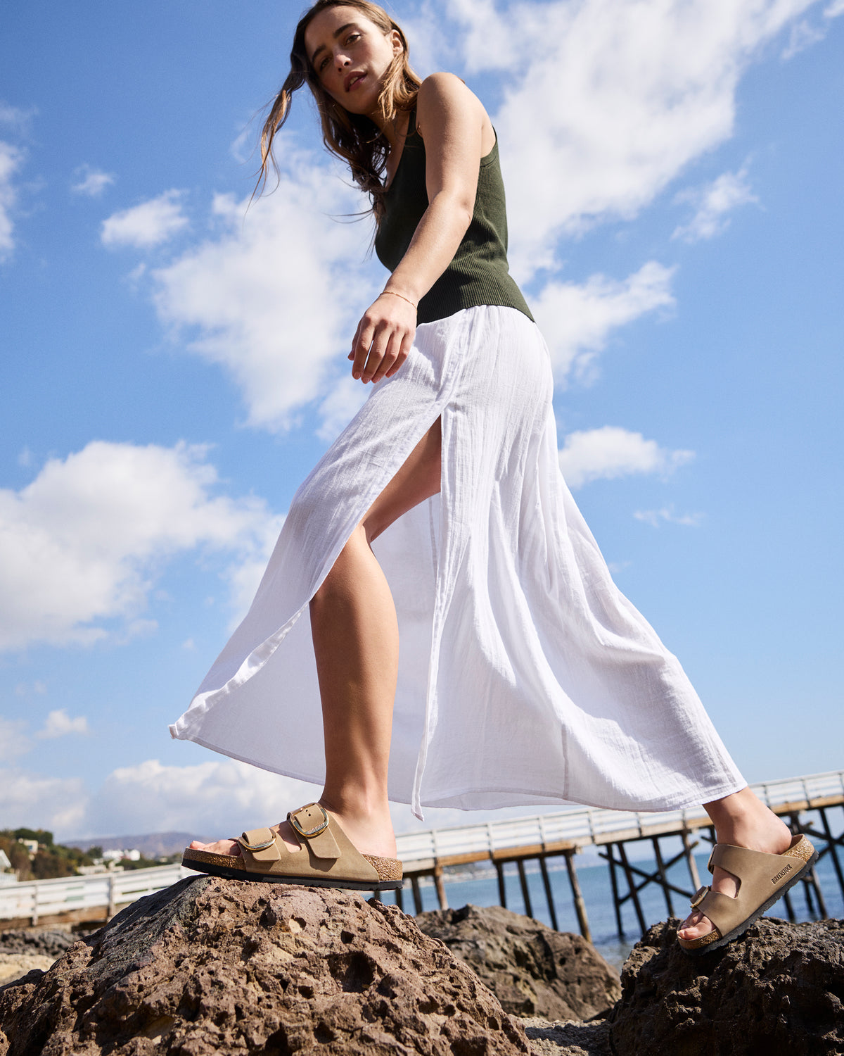 Lifestyle shot of a young woman wearing the Birkenstock Arizona Big Buckle Sandal in Tobacco Brown Oiled Leather on a rock surface at a beach setting wearing a soft linen skirt and dark green tank.