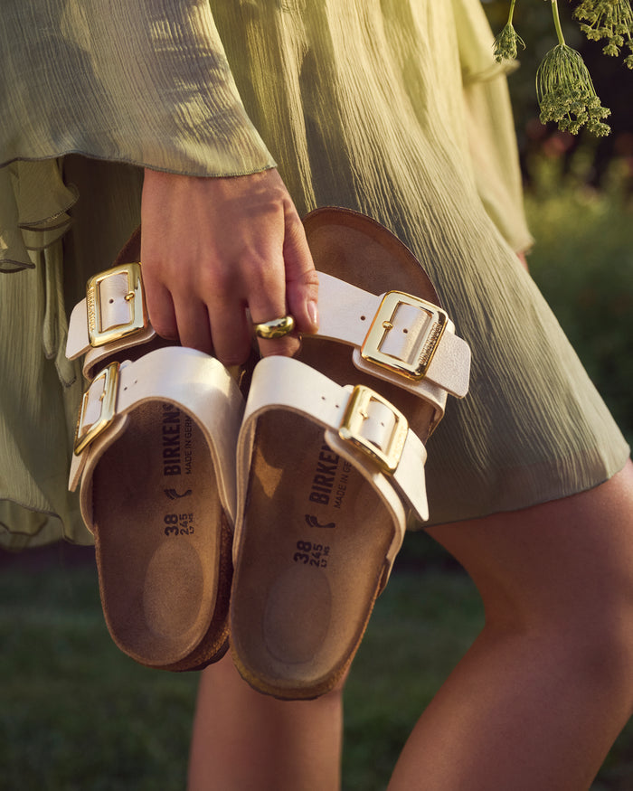 Person holding Birkenstock Sydney Luxe Buckle sandals in Graceful Pearl White with gold buckles in a natural setting