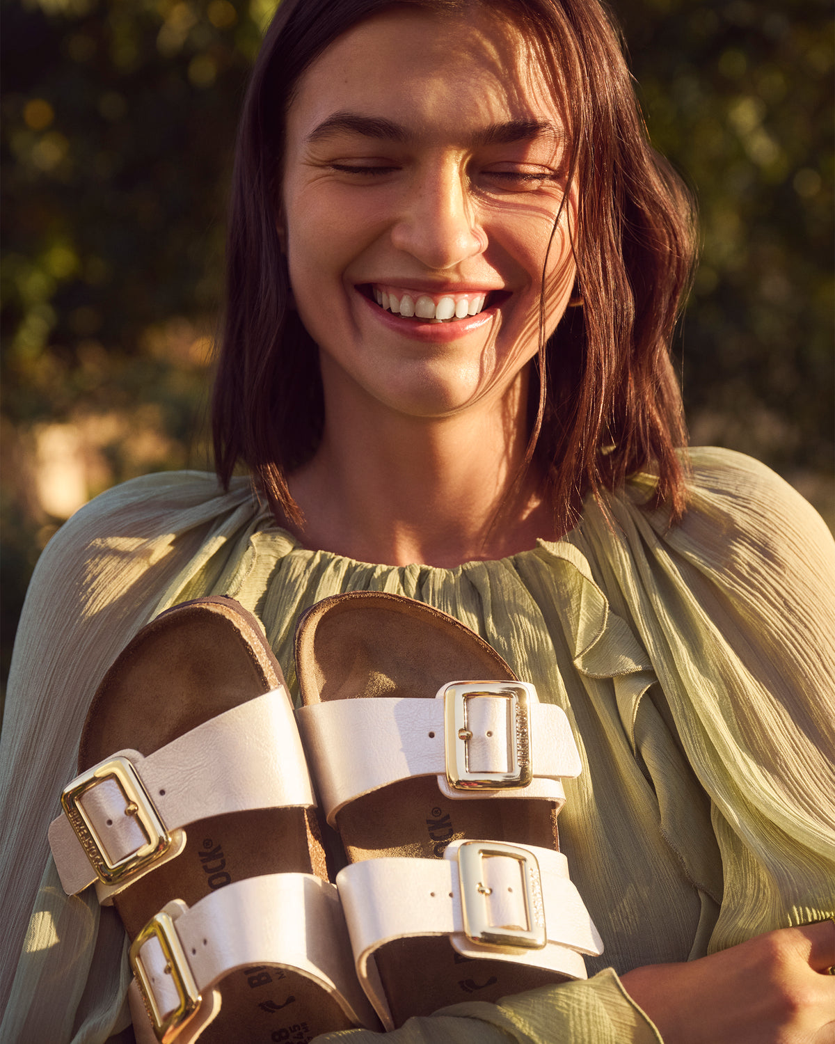 Woman holding a pair of pearl white Birkenstock Sydney Luxe Buckle sandals with gold buckles outdoors