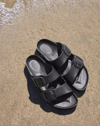 Pair of black EVA Birkenstock sandals with Big Buckle Stealth detailing in a sandy beach setting