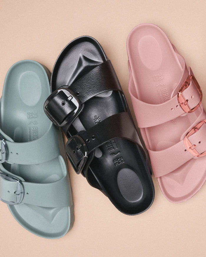 Three Birkenstock Arizona Big Buckle Sandals with two-strap design and similarly-colored translucent buckles on a beige background.