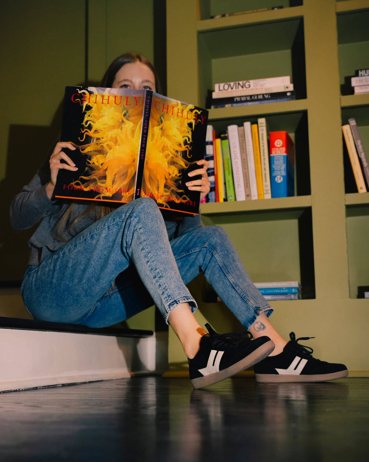 Person sitting on a step reading a book with a vibrant cover in a room with a bookshelf wearing the women's Gabor City Sneaker 73.300.17 in Black