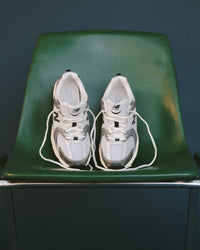 Pair of white sneakers with black accents on a green chair against a dark background. New Balance 530 Sneaker in Silver Metallic with Black Cement