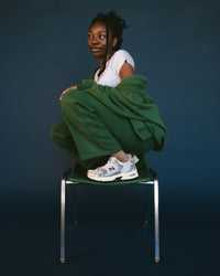 Person sitting on a chair wearing green pants and a white shirt against a dark background. New Balance 530 Sneaker in Silver Metallic with Black Cement