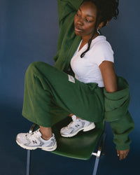 Person wearing a green outfit with New Balance 530 Sneaker in Silver Metallic with Black Cement sneakers sitting on a chair against a blue background