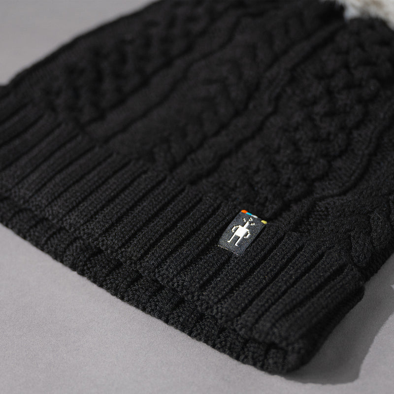 Logo patch detail view of the Women's Smartwool Lodge Girl Soft Merino Knit Beanie with Faux Fur Pom in Black and Gray