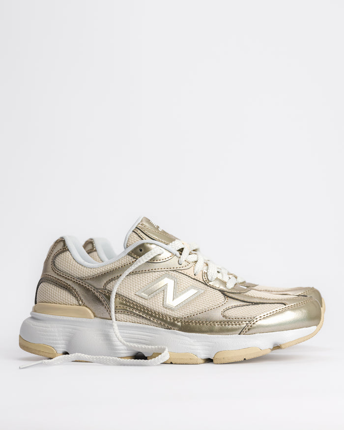 A New Balance 603 sneaker in metallic gold and beige with white laces and a chunky white and beige sole, shown from a side profile on a light gray background. W60347S 