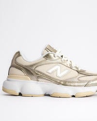 A New Balance 603 sneaker in metallic gold and beige with white laces and a chunky white and beige sole, shown from a side profile on a light gray background. W60347S 