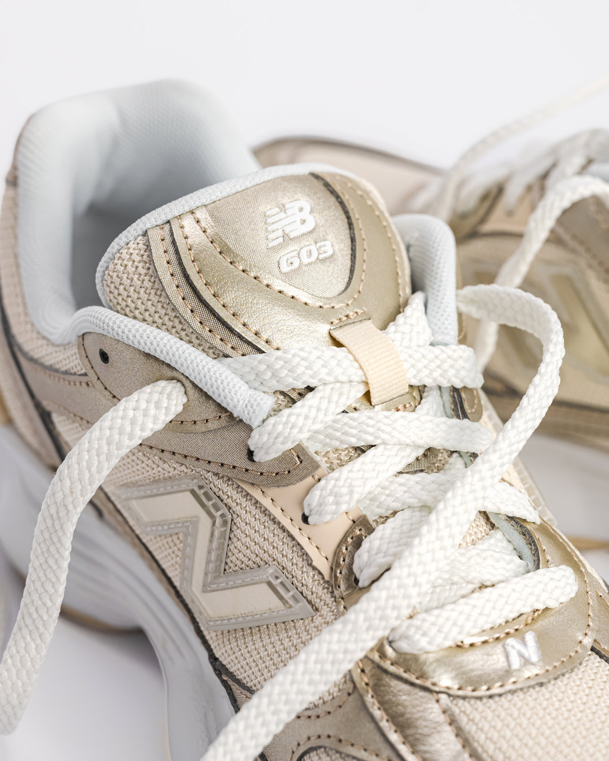 A New Balance 603 sneaker in metallic gold and beige with white laces and a chunky white and beige sole, shown from a side profile on a light gray background. W60347S 