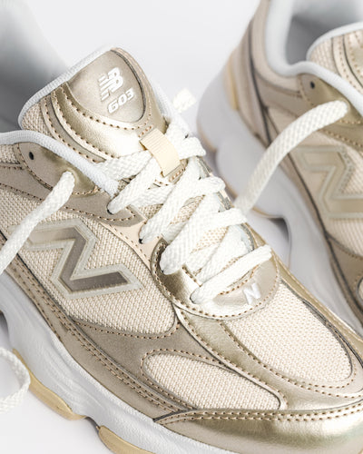 A New Balance 603 sneaker in metallic gold and beige with white laces and a chunky white and beige sole, shown from a side profile on a light gray background. W60347S 